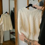 Woman holding and trying on a knitted sweater while observing her reflection in a mirror, showcasing fashion styling.